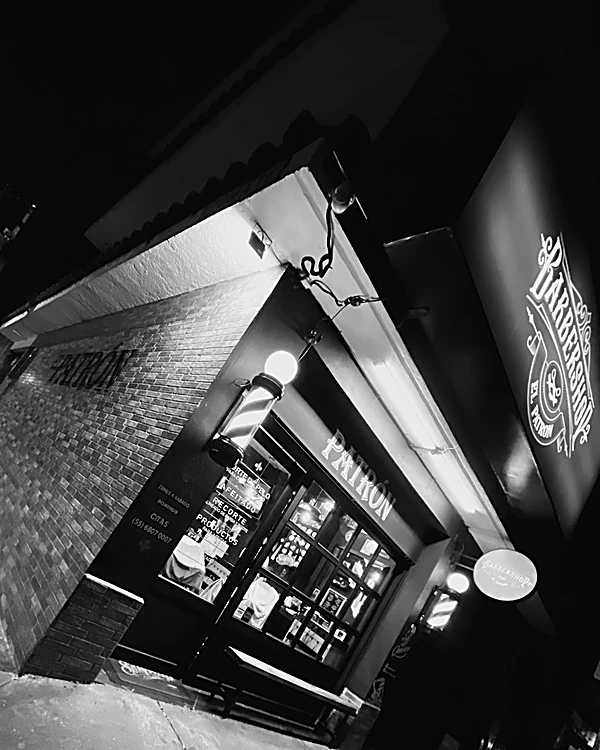 La esquina de noche en blanco y negro: muro de ladrillo con la palabra PATRÓN, poste de barbero encendido y el escudo iluminado Barbershop El Patrón