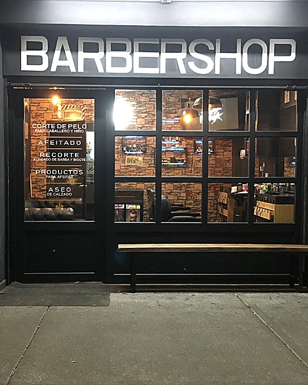 Fachada de El Patrón de noche: letrero BARBERSHOP en blanco sobre negro, el ventanal cuadriculado y, en el vidrio de la puerta, la lista de servicios