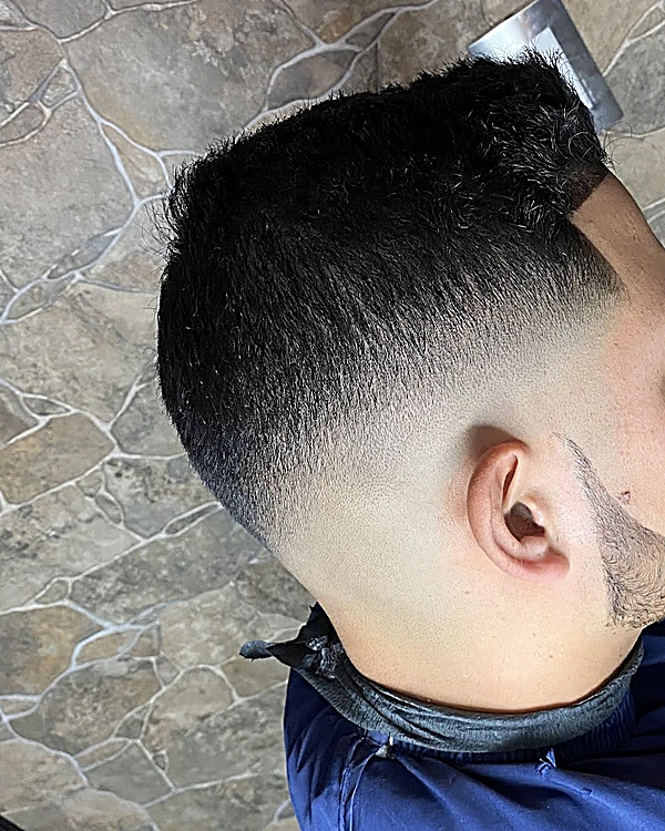 Fade bajo con el cabello corto arriba y la barba delineada en línea fina, de perfil, con capa azul sobre el piso de piedra