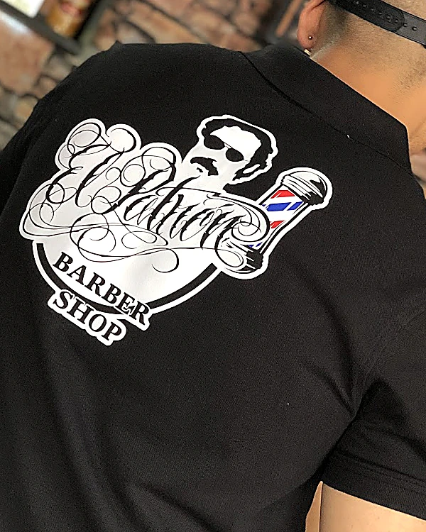 Espalda del polo negro del equipo con el logo de El Patrón: un barbero de lentes oscuros y bigote, la palabra Patrón en caligrafía, el poste y Barber Shop