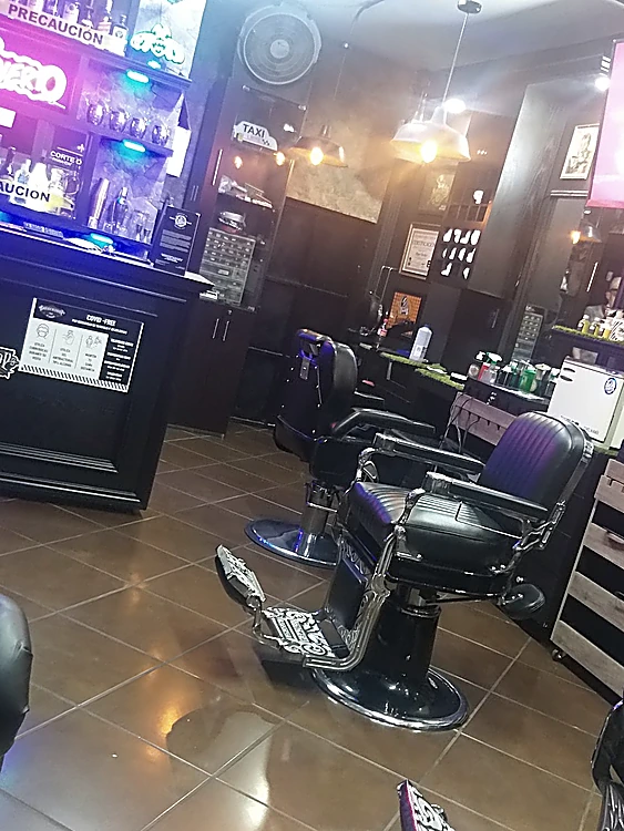 Dos sillas de barbero negras con respaldo cromado ornamentado, la barra con luces de neón y el piso de loseta